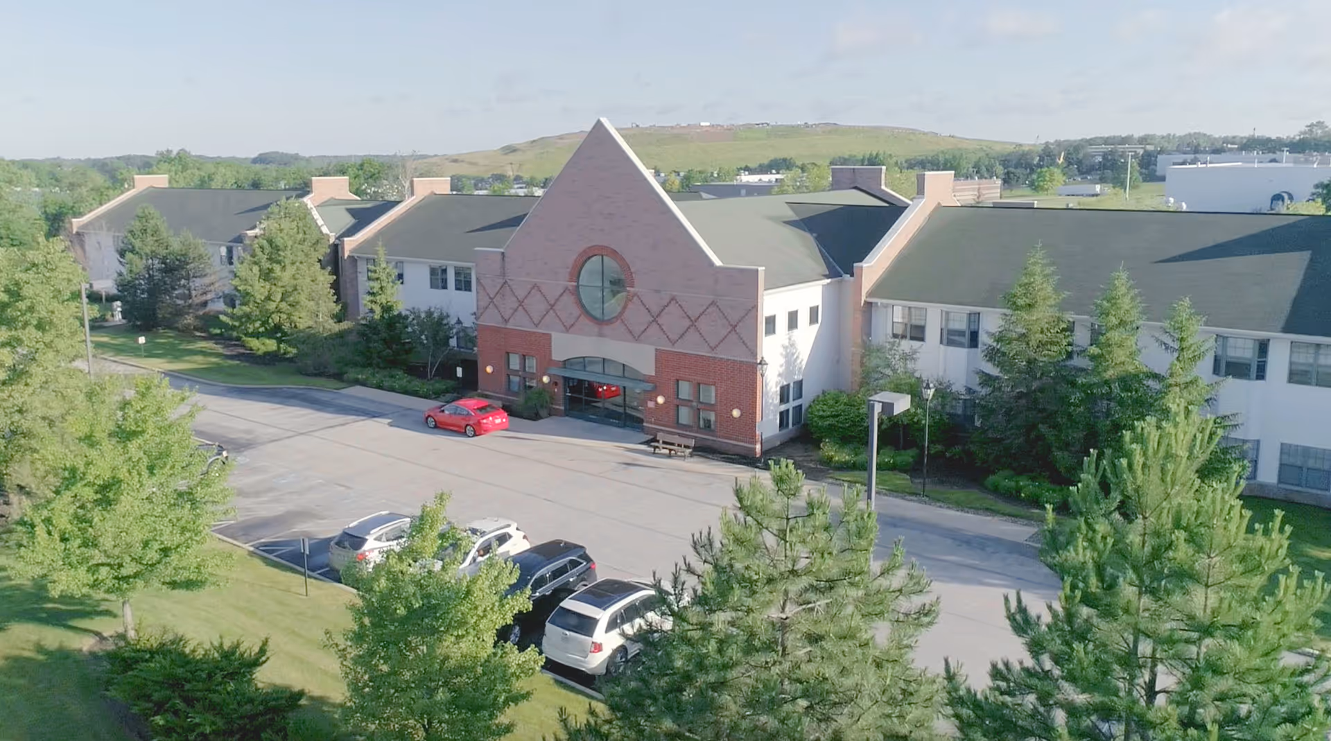 Exterior view of Stratford Commons Assisted Living facility showing a large building with a peaked roof entrance, surrounded by trees and a parking lot with several cars parked.