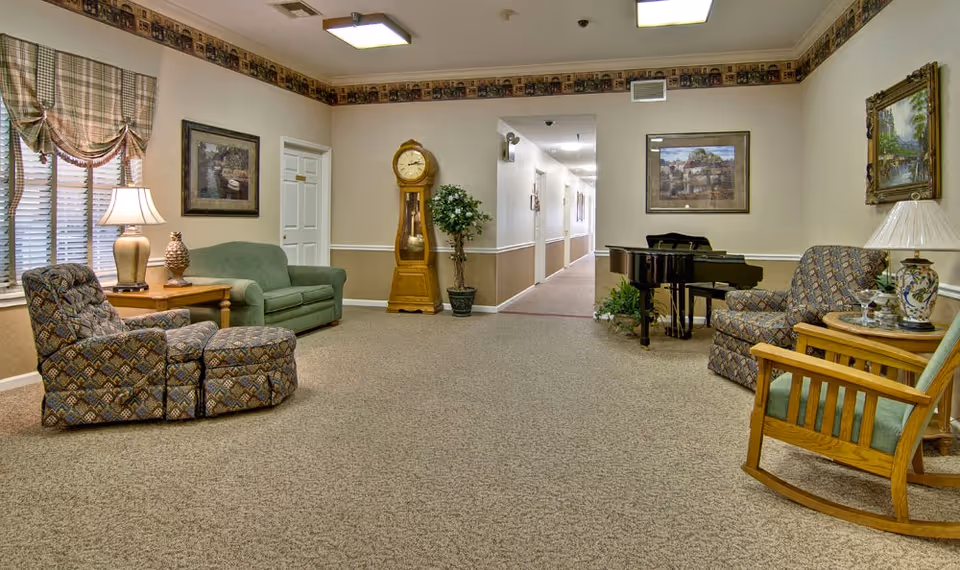 A cozy senior living facility common area with patterned armchairs, a green loveseat, a wooden rocking chair, side tables with lamps, a grandfather clock, potted plants, framed paintings on the walls, and a black grand piano near a hallway.
