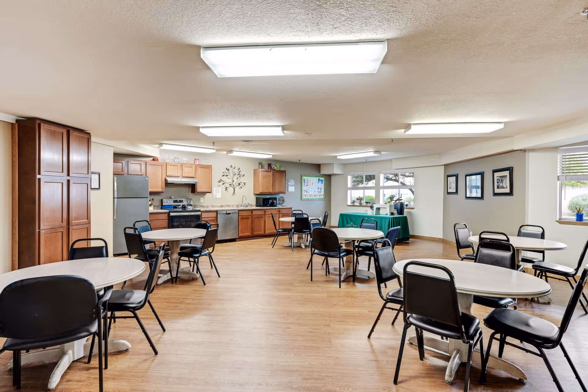 A spacious dining area with several round tables and black chairs arranged around them. The room has wooden flooring and a kitchen area with wooden cabinets, a refrigerator, stove, and dishwasher along one wall. There are large windows letting in natural light, framed pictures on the walls, and fluorescent ceiling lights illuminating the space.