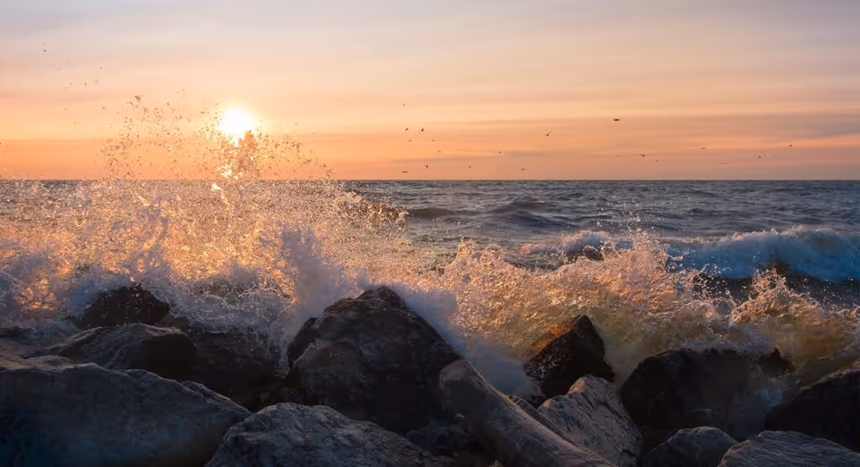 Waves crashing against a rocky shoreline at sunset with the sun low on the horizon.
