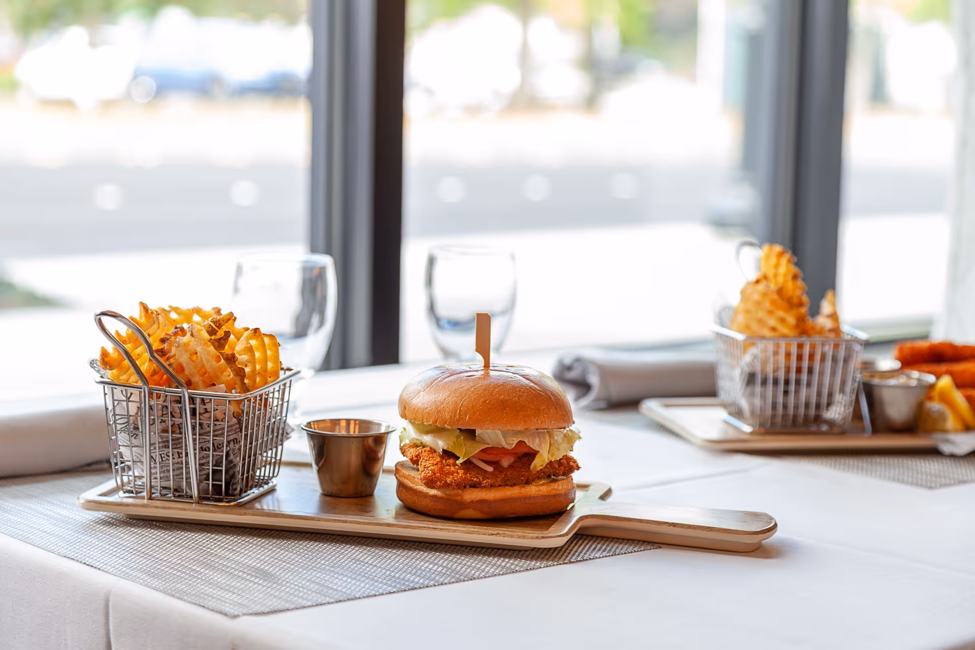 A fried chicken sandwich with lettuce and a side of waffle fries served on wooden boards on a table by a window.