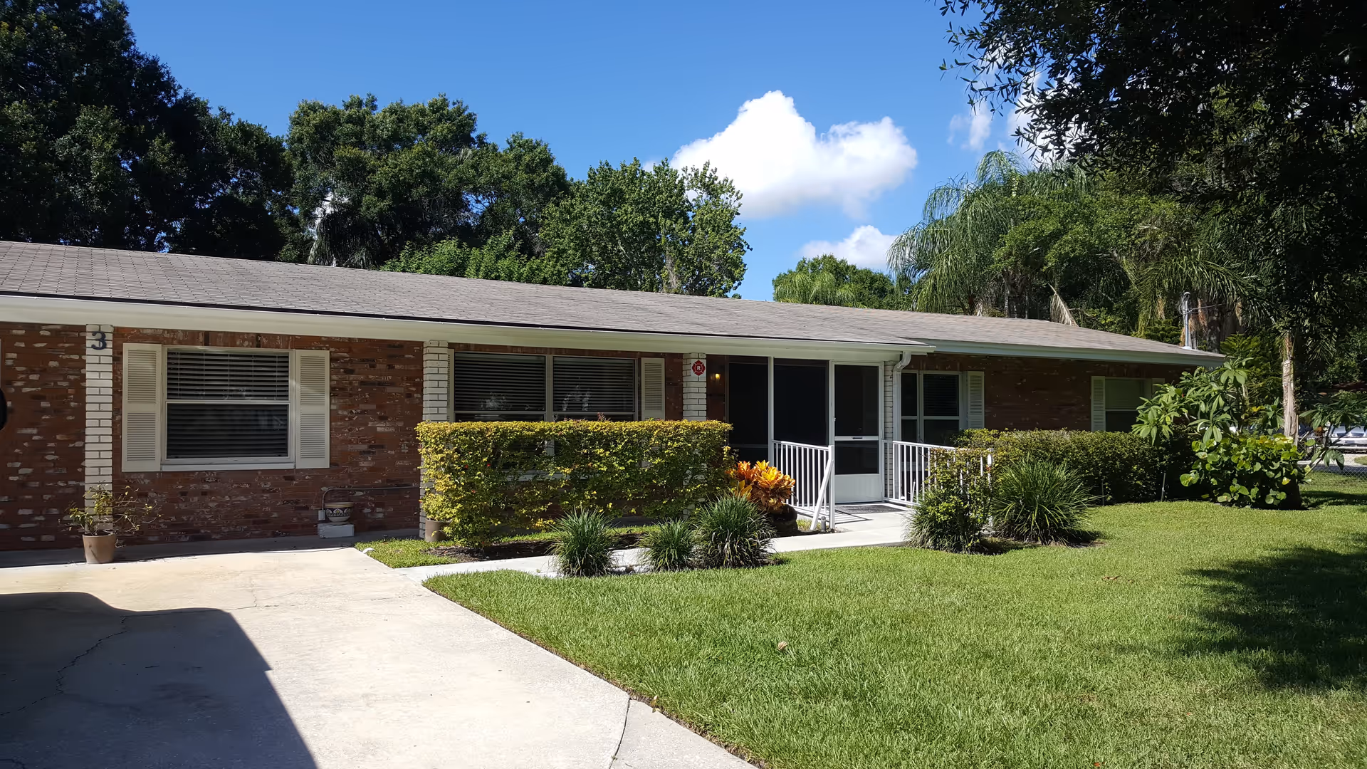 Single-story brick building with white trim and a gray shingled roof, surrounded by green grass, bushes, and trees under a clear blue sky with a few clouds. There is a concrete driveway and a wheelchair accessible ramp leading to the entrance.