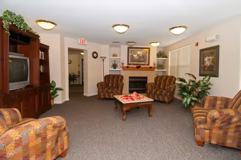 A cozy living room area with patterned armchairs arranged around a wooden coffee table. The room features a fireplace with decorative items on the mantel, framed artwork on the walls, a wooden entertainment center with a television, and several potted plants. The ceiling has multiple round light fixtures, and there is an exit sign above a doorway leading to another room.