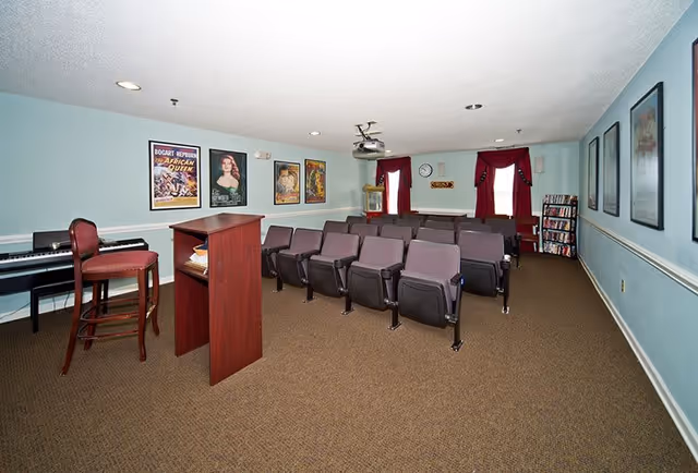 A small theater or presentation room with rows of purple cushioned chairs facing a wooden podium. The walls are light blue with framed vintage movie posters. There is a piano with a tall chair on the left side, a popcorn machine near the back, and a bookshelf with DVDs or books. Two windows with red curtains are at the back of the room.