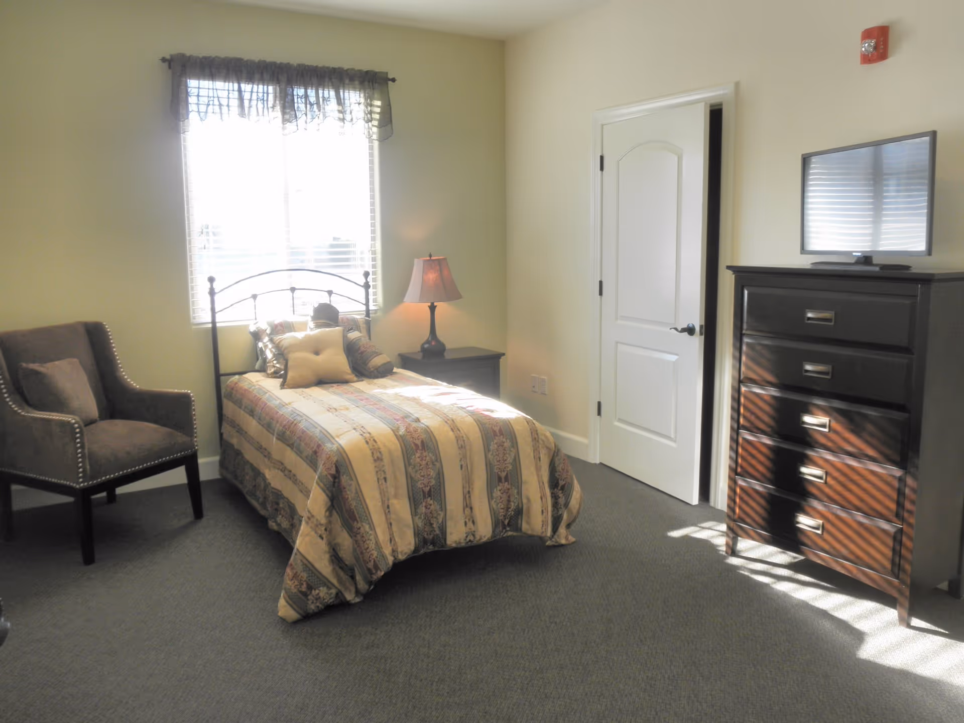 A bedroom with a single bed covered in a patterned bedspread, a cushioned armchair to the left, a nightstand with a lamp next to the bed, a window with sheer curtains letting in sunlight, and a dark wooden dresser with a flat-screen TV on top.