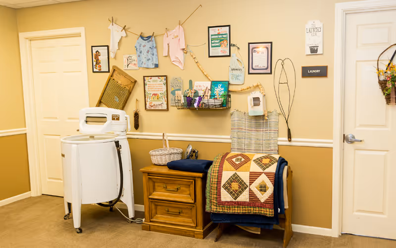 A cozy laundry room with vintage decor including an old-fashioned white washing machine, a wooden side table with a basket, a quilt draped over a chair, and various framed pictures and laundry-themed decorations on the beige walls. There are two white doors, one labeled 'Laundry'.