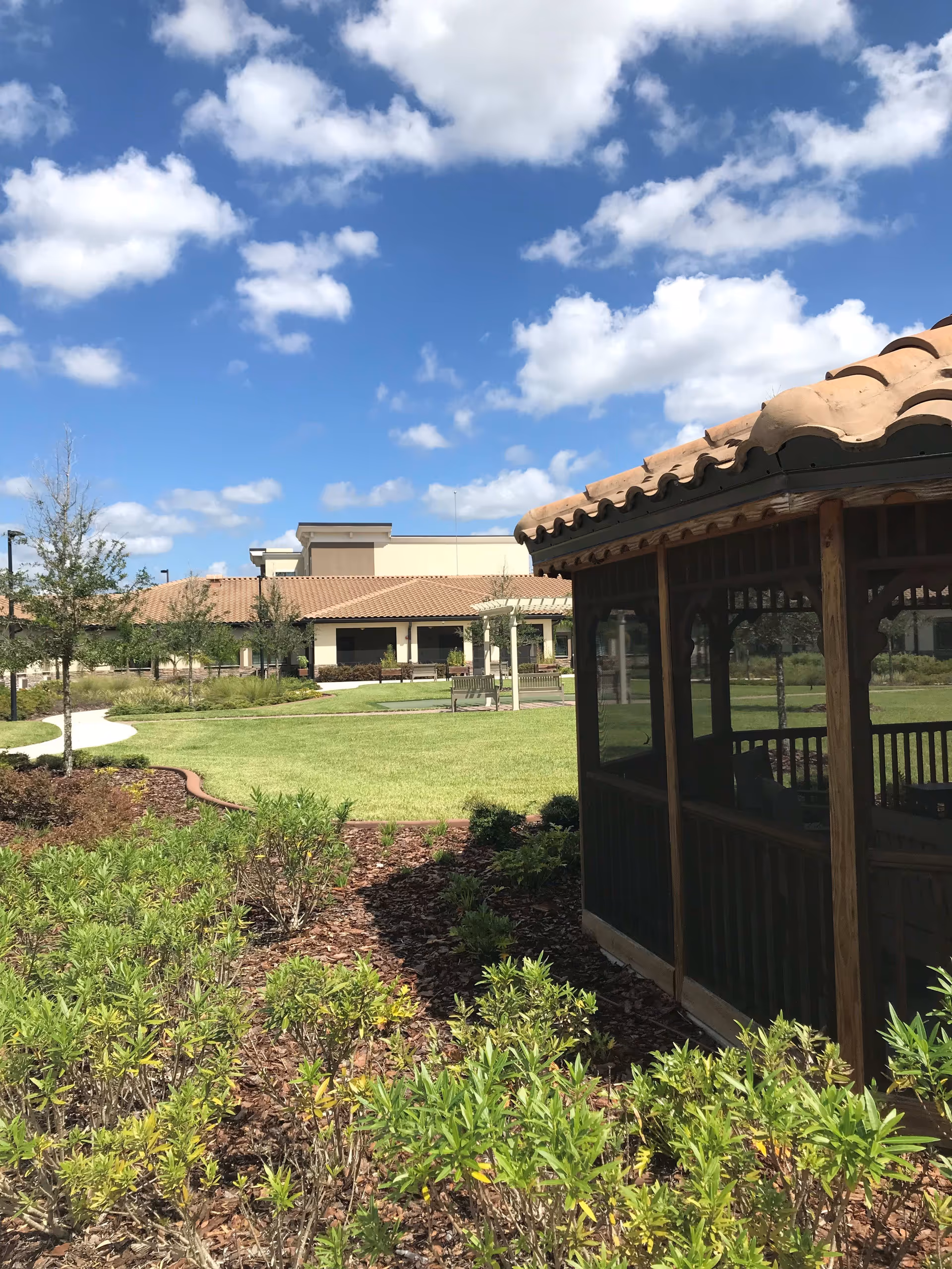 Outdoor garden area at Discovery Village At Deerwood Assisted Living & Memory Care featuring a wooden gazebo with a tiled roof on the right, green shrubs and plants in the foreground, a well-maintained lawn, benches, and a building with a tiled roof in the background under a partly cloudy blue sky.