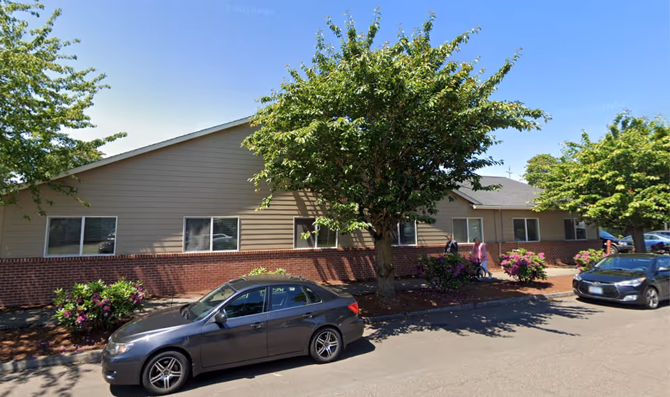 Exterior view of a single-story senior living facility building with beige siding and a brick lower facade. There are several windows along the building, a large tree, and some bushes in front. Two cars are parked on the street, and two people are walking on the sidewalk near the entrance.