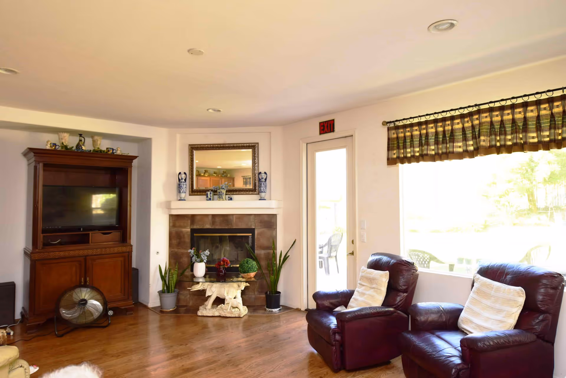 A cozy living room with two dark brown leather recliners with light-colored pillows, a fireplace with decorative vases and plants on the mantel, a wooden TV cabinet with a flat-screen TV, a small table with plants in front of the fireplace, and a large window with striped curtains letting in natural light. There is a door with an exit sign above it leading outside.