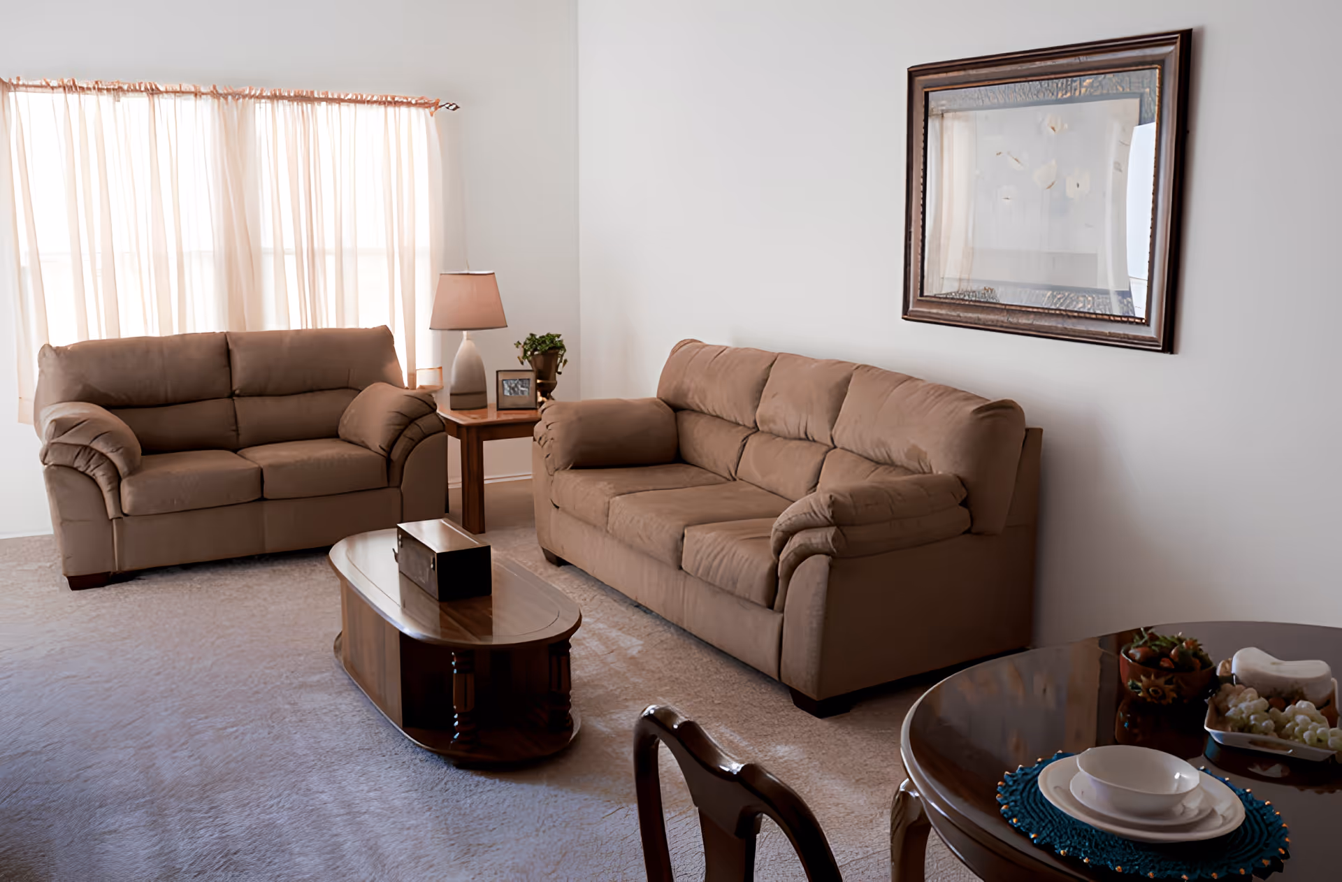 Sunlit living room with two beige sofas, a wooden coffee table and side table with a lamp, and a small dining table with place settings.