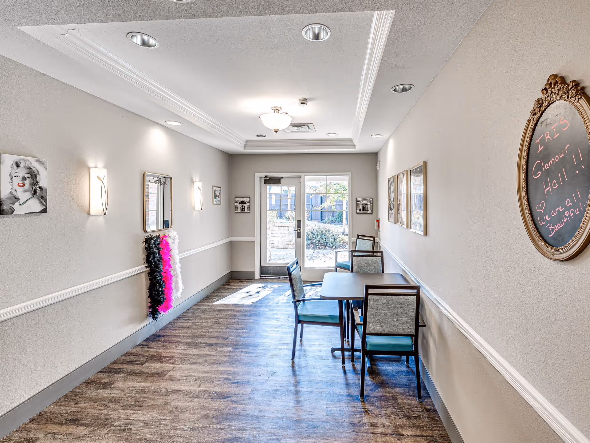 A bright hallway in a senior living facility with wood flooring, beige walls, and a white ceiling with recessed lighting. On the right wall, there is an oval chalkboard with the message 'IRIS Glamour Hall!! We are all Beautiful' written in pink chalk. Below the chalkboard are three framed pictures. On the left wall, there is a black and white portrait of Marilyn Monroe, a mirror, and colorful feather boas hanging beneath the mirror. At the end of the hallway, there is a glass door leading outside, with two tables and chairs arranged near the door.