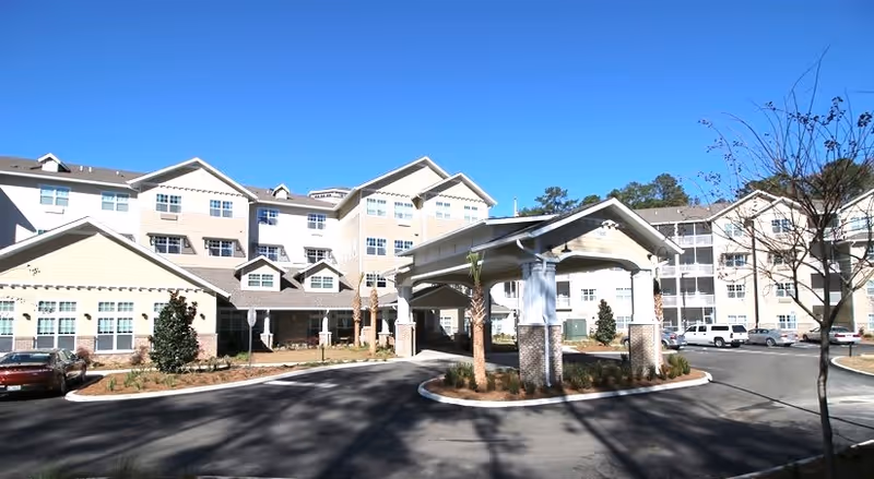 Exterior view of a multi-story senior living facility with beige siding and white trim, featuring a covered entrance driveway with columns and landscaped areas with small trees and shrubs under a clear blue sky.