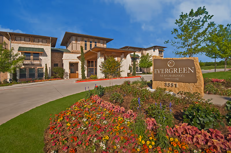 Front exterior of the Evergreen at Richardson senior living building with a stone entrance sign and colorful landscaped flowerbeds.