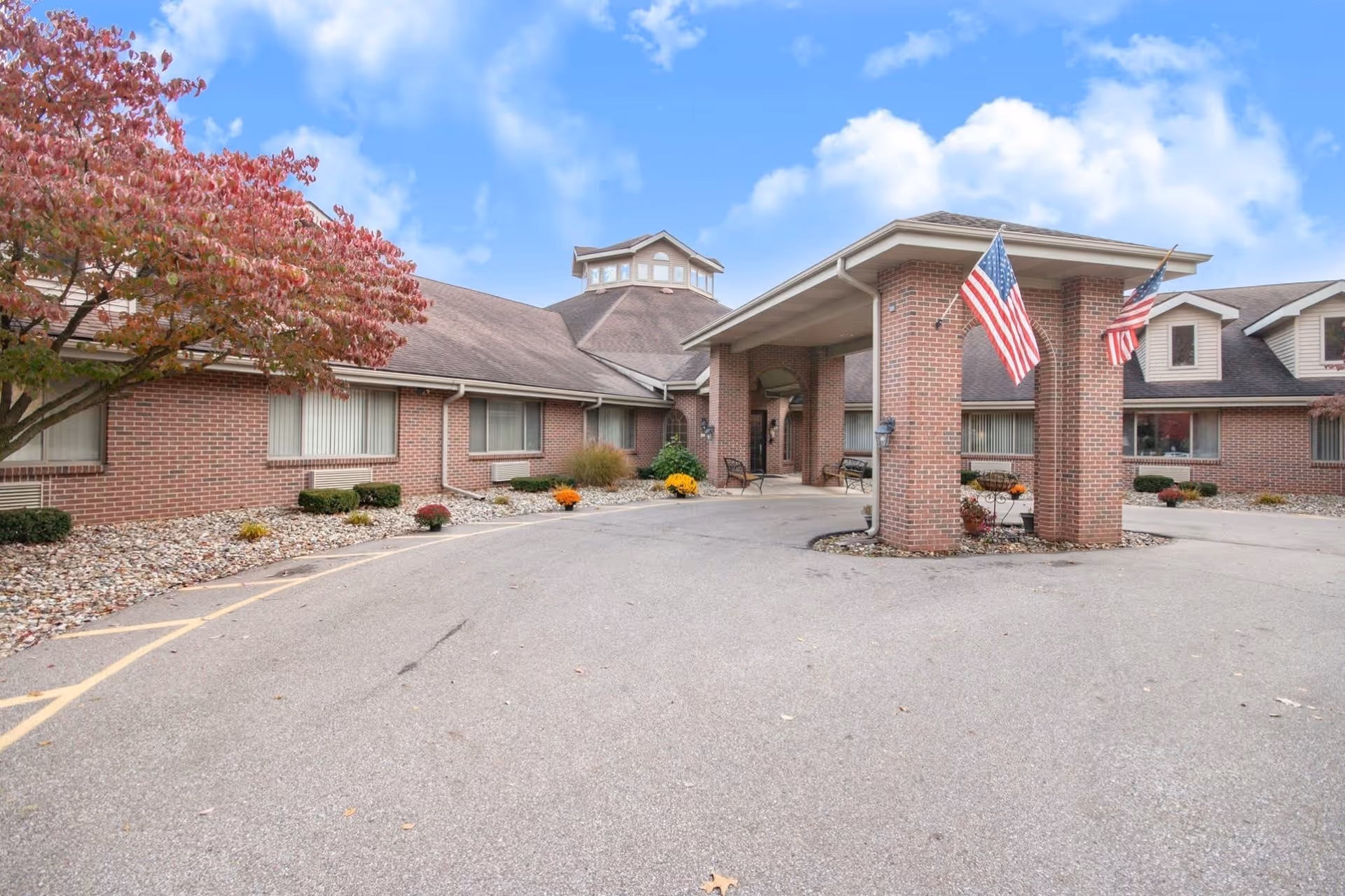 Exterior view of Brentwood at Elkhart Assisted Living facility showing a brick building with a covered entrance supported by brick columns, two American flags, a driveway, and landscaping with small bushes and a tree with red leaves under a partly cloudy sky.