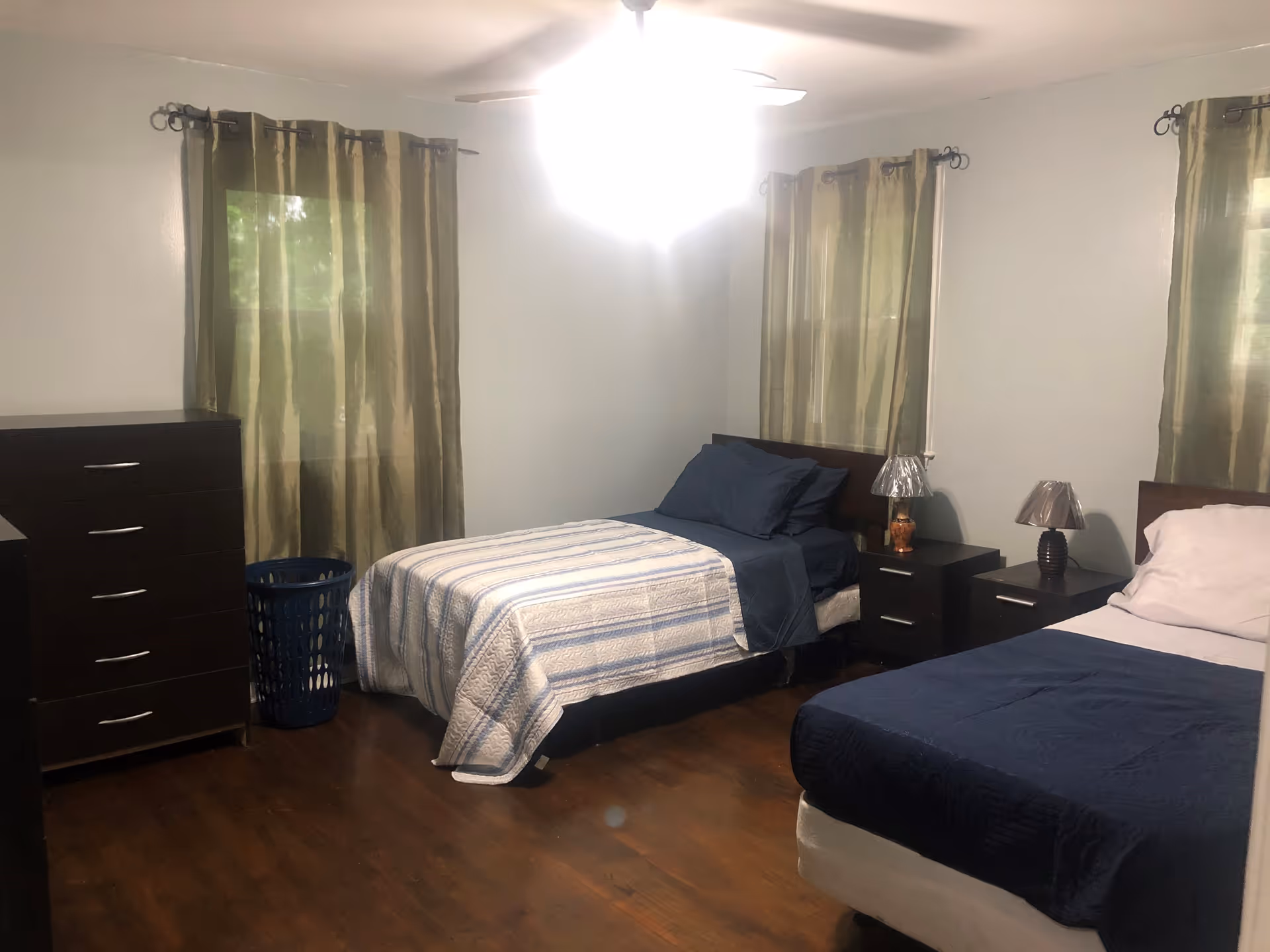 Two twin beds with blue quilts, nightstands, a dresser, a laundry basket, and curtains.