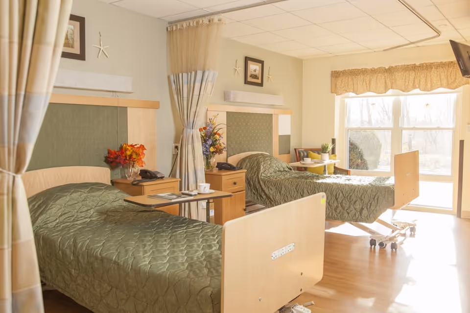 A bright and clean room with two hospital-style beds covered in green quilted bedspreads. Each bed has a wooden headboard and footboard, a bedside table with a telephone and flowers, and a curtain that can be drawn for privacy. Large windows let in natural light, and there is a TV mounted on the wall.