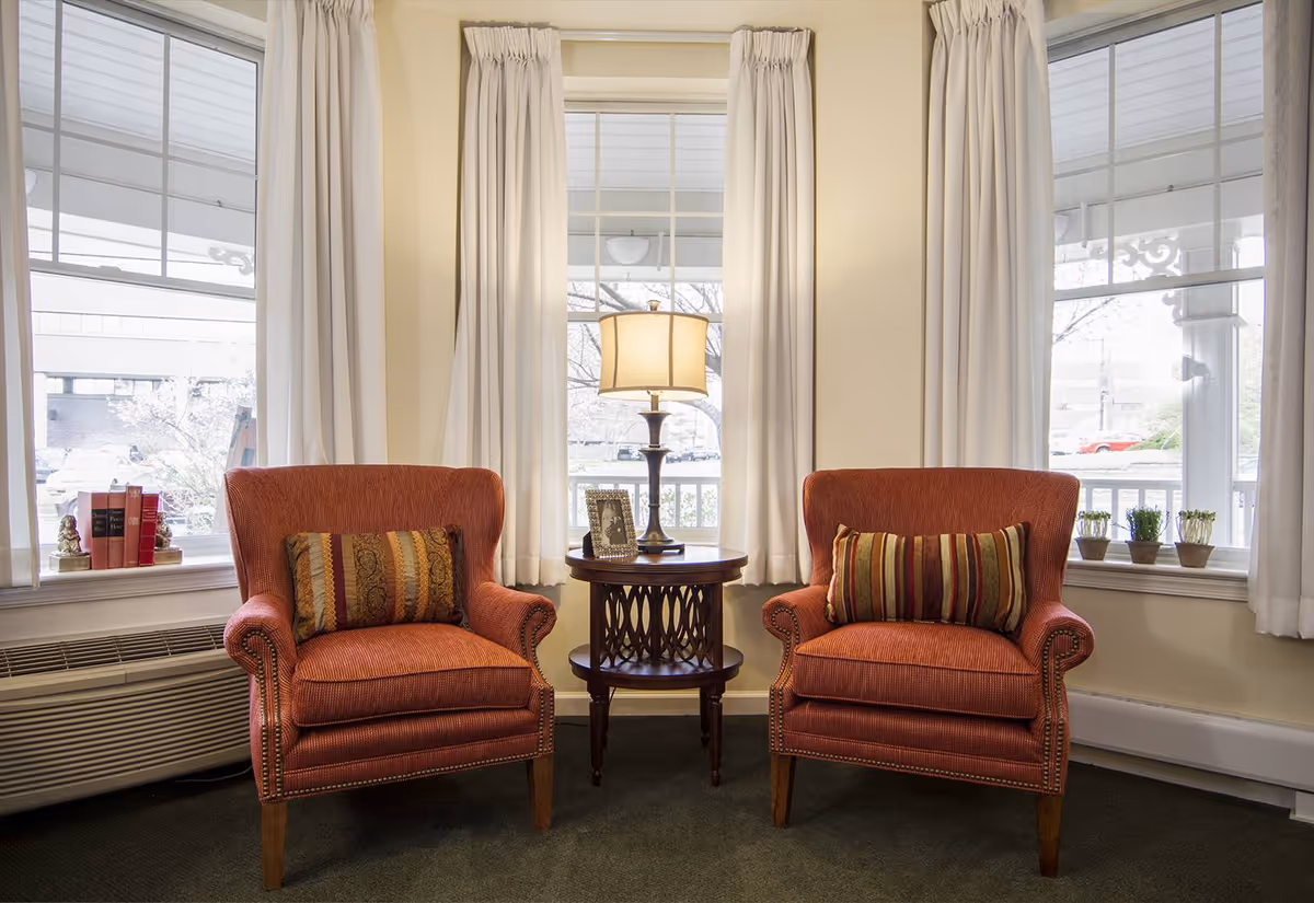 Two orange upholstered armchairs with patterned cushions are placed on either side of a round wooden side table with a lamp and a framed photo. The setting is in front of three large windows with white curtains, and small potted plants and books are on the windowsills.