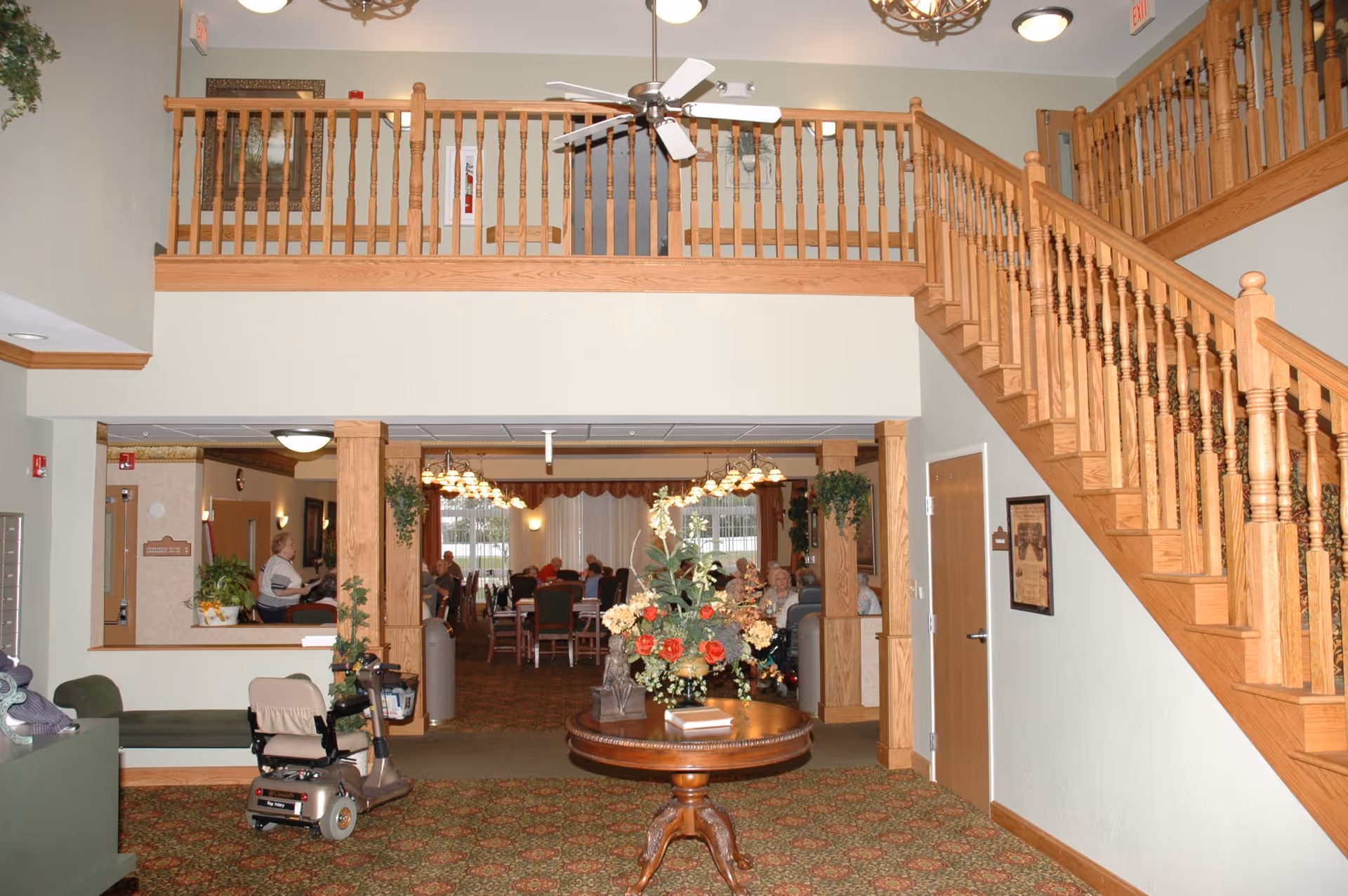 Interior view of a senior living facility showing a carpeted area with a wooden staircase on the right and a round wooden table with a floral arrangement in the center. In the background, there is a dining area with several elderly people seated at tables under hanging light fixtures. The upper level has a wooden railing and a ceiling fan.