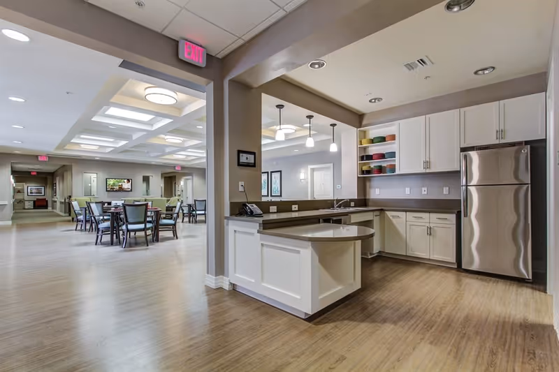 Open communal kitchen with an island, stainless refrigerator, and adjacent dining area with tables and chairs.