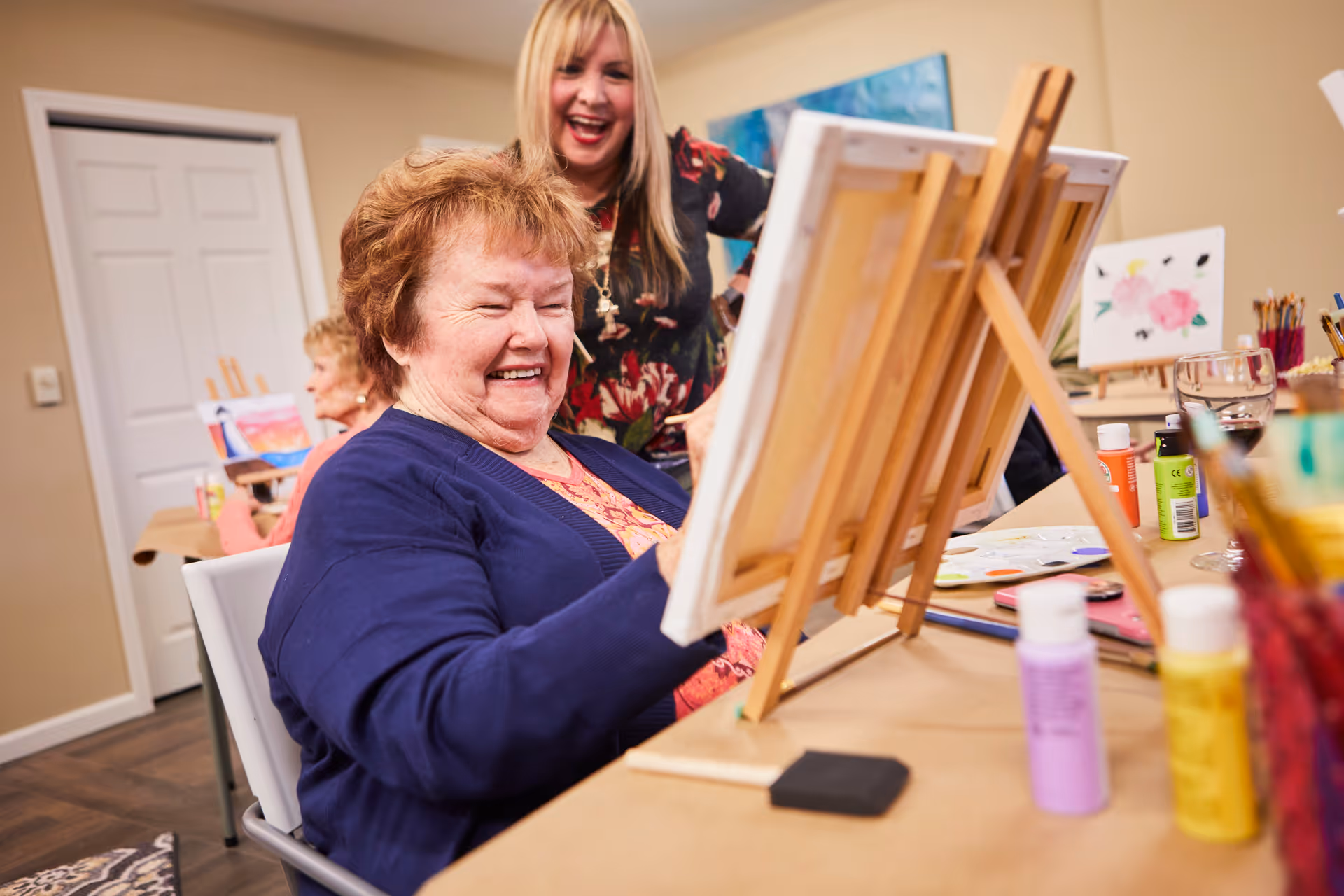 An elderly woman smiling and painting on a canvas set on an easel in a room, with a younger woman standing behind her smiling. There are art supplies and paint bottles on the table, and another elderly woman painting in the background.