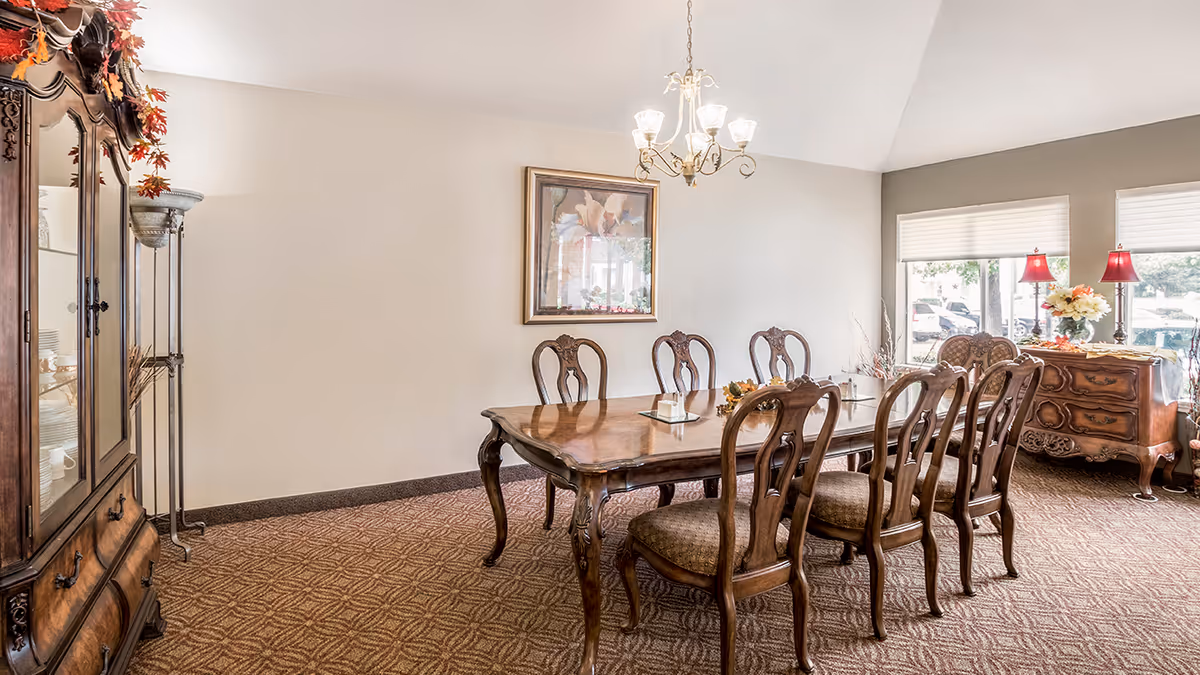 A well-lit dining room with a large wooden dining table surrounded by eight matching wooden chairs with cushioned seats. The room features a chandelier hanging above the table, a framed floral painting on the wall, a wooden cabinet with glass doors on the left, and a wooden sideboard with two red lamps and a flower arrangement near the windows on the right. The carpet has a patterned design, and the windows let in natural light.