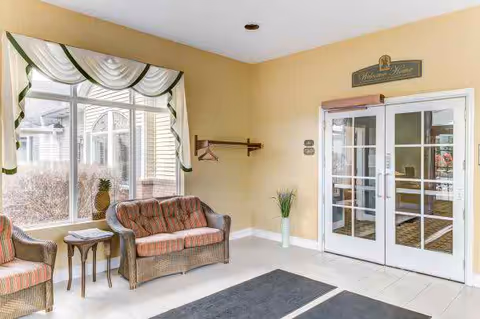 A bright and welcoming interior space with beige walls and white tiled floor. There are two wicker chairs with cushions and a small wooden side table near a large window with white and green curtains. A coat rack is mounted on the wall, and a tall vase with green plants stands near double glass doors. Above the doors is a sign that reads 'Walden Place'.