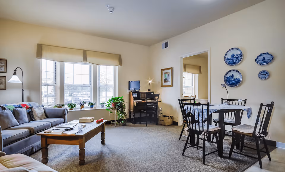 A bright living room with a gray sofa, wooden coffee table, and a small dining table with four chairs. There are large windows with beige valances letting in natural light. A small desk with a computer and lamp is in the corner, and decorative blue plates hang on the wall.