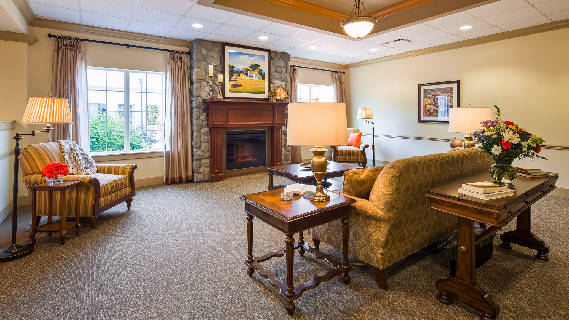 A cozy living room area with a patterned sofa, two armchairs, wooden side tables with lamps, a stone fireplace with a wooden mantle, and framed artwork on the walls. There is a large window with curtains letting in natural light and a vase of flowers on a wooden console table behind the sofa.