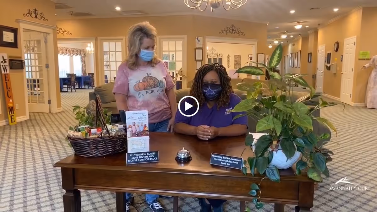 Two women wearing face masks are at a wooden reception desk in a senior living facility lobby. One woman is standing and the other is sitting behind the desk. The desk has a basket with items, informational signs, a service bell, and a potted plant. The background shows a carpeted hallway with doors and framed pictures on the walls.