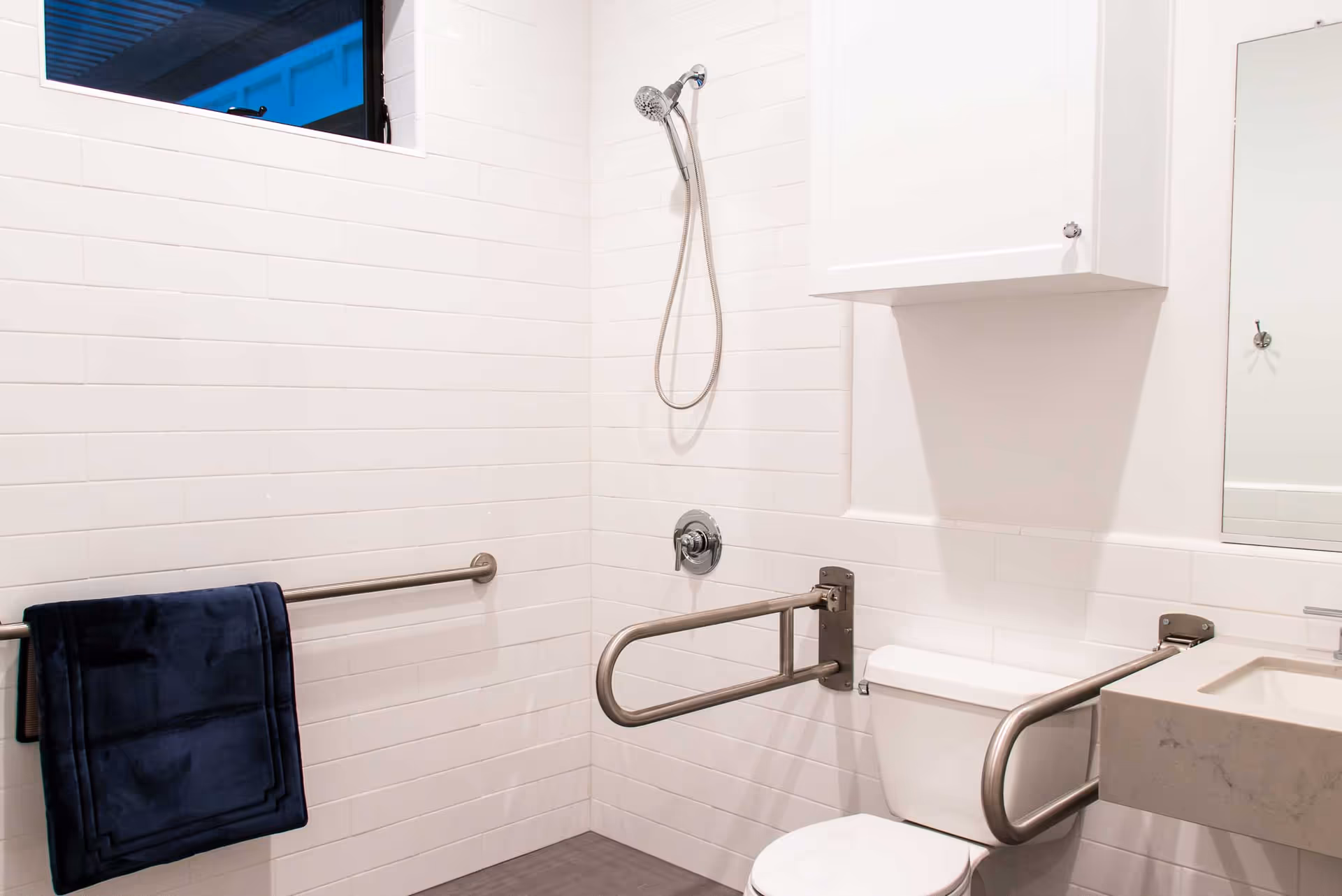 A clean, accessible bathroom with white tiled walls, a showerhead, a toilet with metal grab bars on both sides, a towel rack with a dark blue towel, a white cabinet above the toilet, and a sink with a marble countertop.