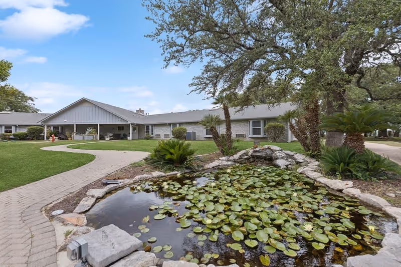 Outdoor garden area at a senior living facility featuring a small pond filled with lily pads and surrounded by rocks, with palm trees and other greenery nearby. A paved walkway curves around the pond leading to a single-story building with a covered porch under a partly cloudy sky.