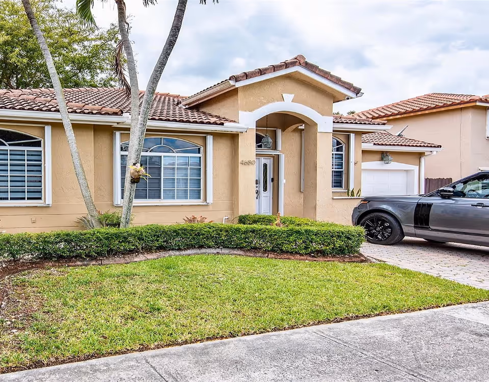 Front exterior of a tan stucco house with an arched entryway, tiled roof, lawn, and a gray SUV parked in the driveway.