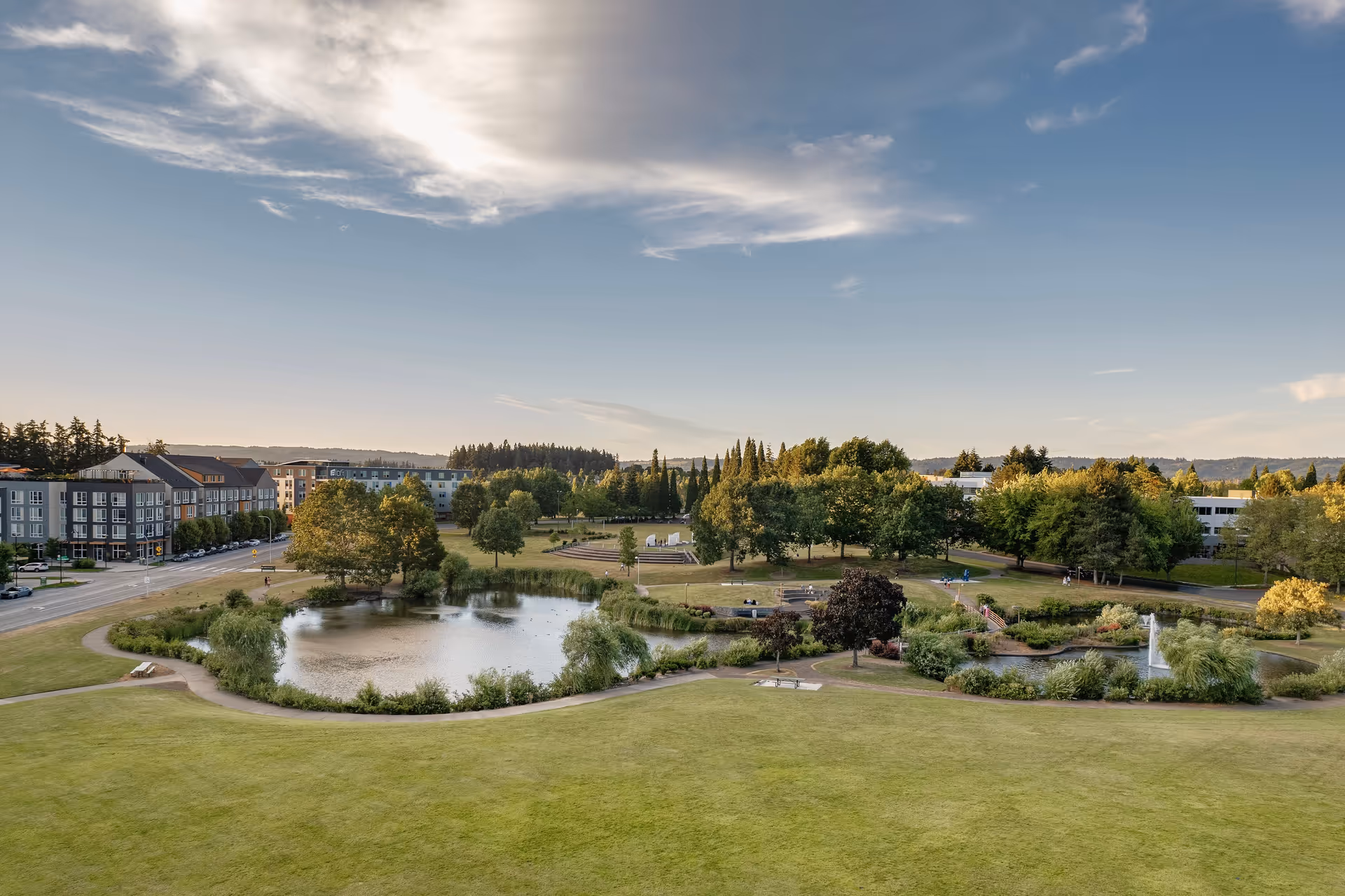 A scenic outdoor park area with a pond, walking paths, trees, and benches under a partly cloudy sky during late afternoon or early evening. Buildings and a road are visible in the background.