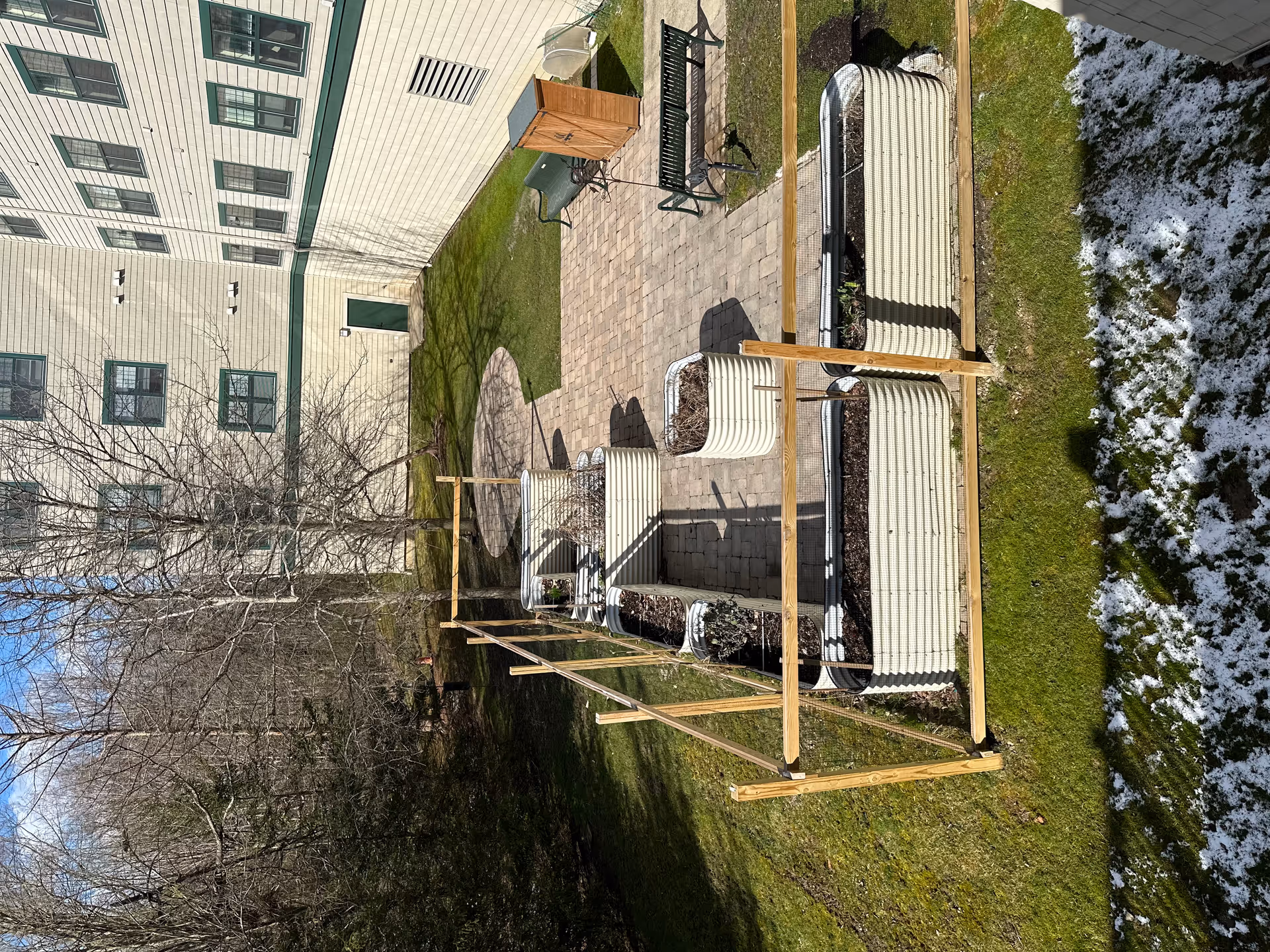 Outdoor garden area at Deer Creek Lodge featuring several raised garden beds enclosed by a wooden frame, a paved patio with benches, a small wooden storage shed, and a building with multiple windows in the background. Some patches of snow are visible on the grass in the foreground.