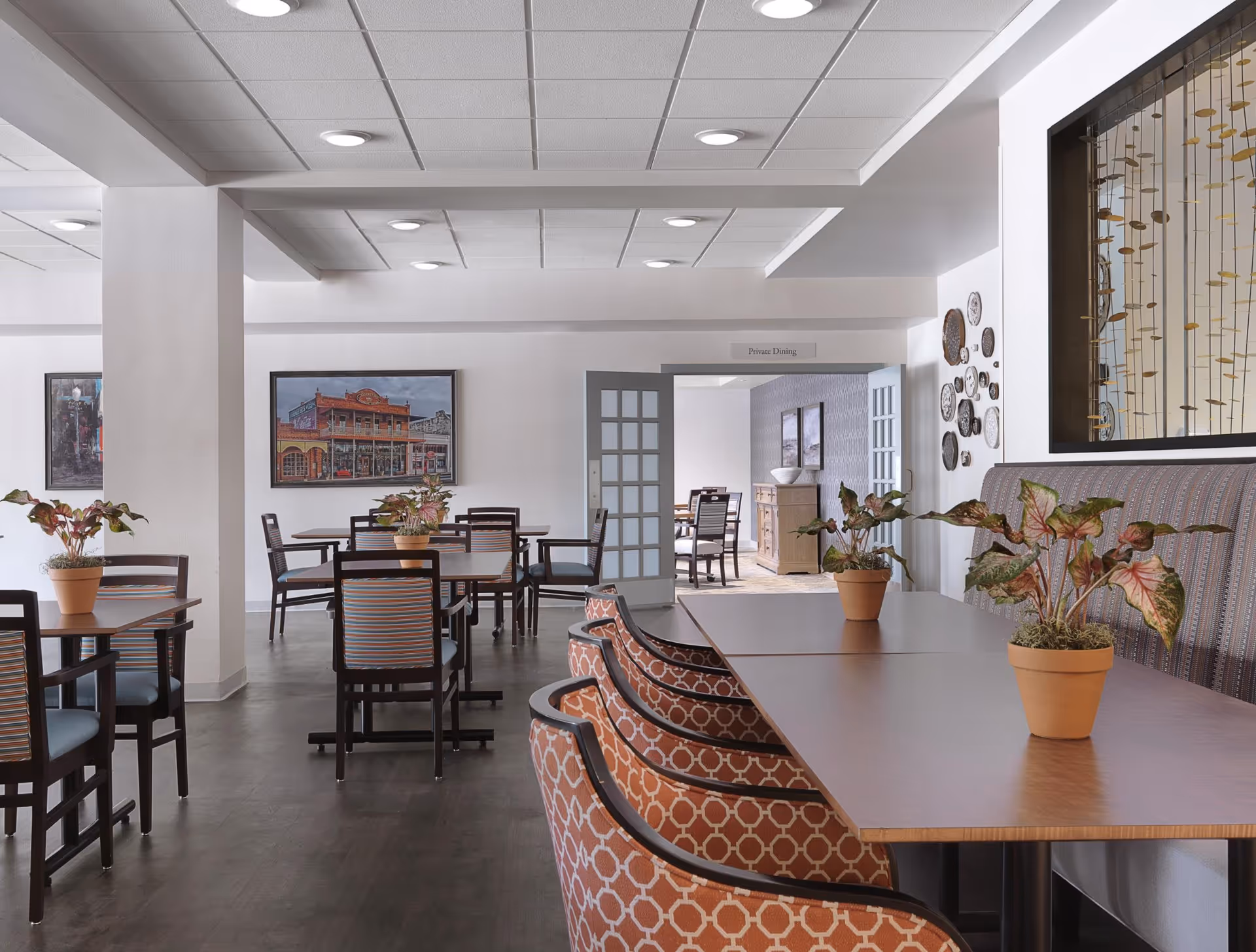 A clean and well-lit dining area in a retirement community with several tables and chairs. The tables have small potted plants as centerpieces. The room features patterned chairs, framed artwork on the walls, and a sign indicating a private dining area through double doors in the background.
