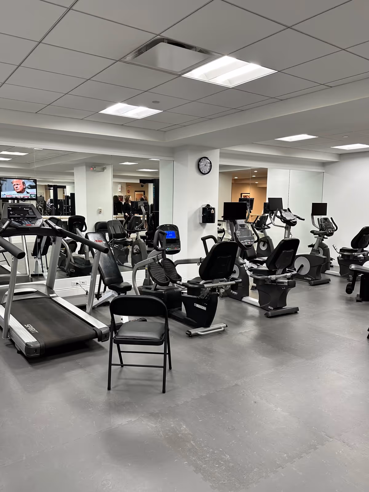 Indoor fitness room with various exercise equipment including treadmills, stationary bikes, and recumbent bikes. There is a black folding chair in the foreground, large mirrors on the walls, and a clock mounted on a white pillar. The room has a gray floor and white ceiling with fluorescent lighting.
