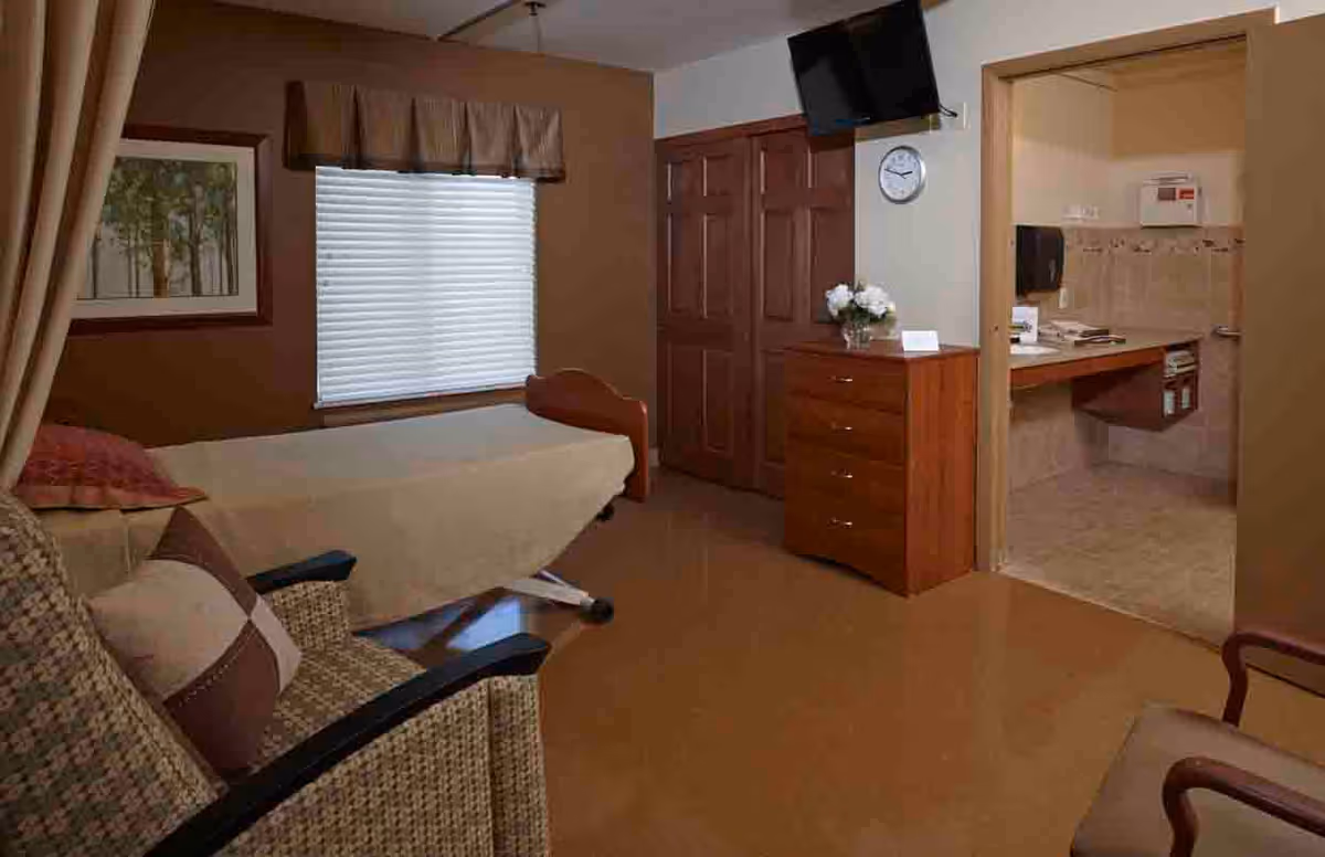 A cozy room with a single bed covered in a beige blanket, a cushioned armchair with a pillow, a wooden dresser with a flower vase, a wall-mounted TV, and a clock. There is an open doorway leading to a small tiled bathroom or wash area with a sink and paper towel dispenser.