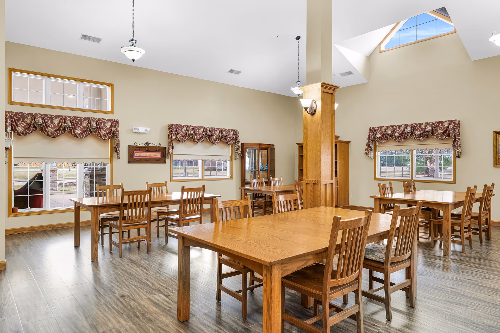 A bright dining room with multiple wooden tables and chairs arranged neatly. The room has large windows with patterned valances, allowing natural light to fill the space. The walls are painted a light beige color and the floor has a wood-like finish. There are ceiling lights and a tall pillar in the center of the room, with a skylight above letting in additional daylight.