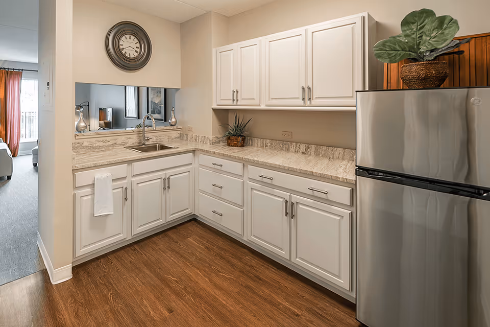 A modern kitchen area with white cabinets and drawers, a stainless steel refrigerator, a sink with a faucet, and a marble countertop. There is a round wall clock above the sink and a potted plant on top of the refrigerator. The kitchen floor is wooden, and an adjacent living room with a window and curtains is partially visible.