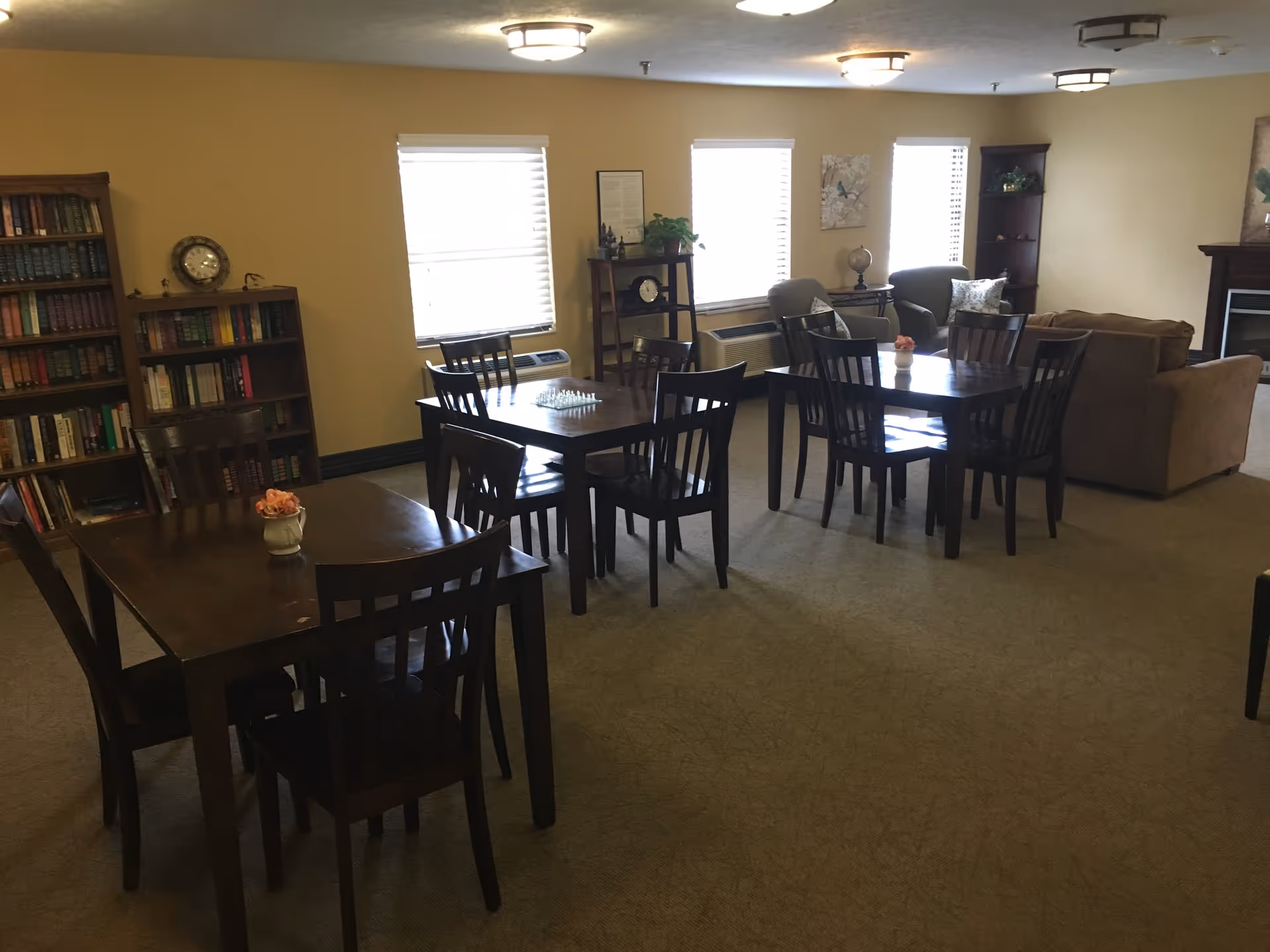 A senior living community common area with several dark wooden tables and chairs arranged for seating. There are bookshelves filled with books along the left wall, and three windows with blinds letting in natural light. The room also features two armchairs, a small table with a globe, a fireplace, and a couch. The walls are painted beige and the carpet is light brown.