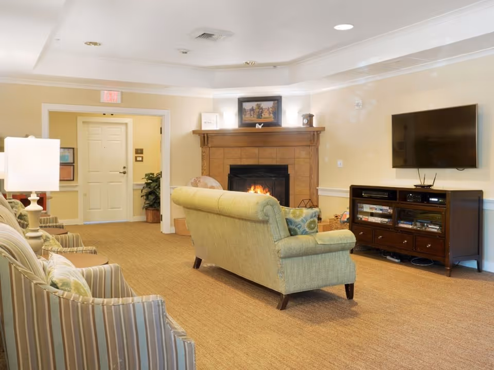Common area living room with a sofa facing a lit fireplace and wall-mounted TV, with striped armchairs and a TV console.
