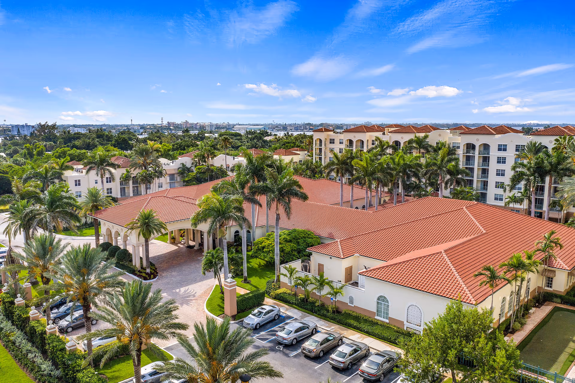 Aerial view of The Residences at The Carlisle Palm Beach showing a large building with red-tiled roofs surrounded by palm trees and greenery. Several parked cars are visible in the parking lot in front of the building, with a clear blue sky overhead.