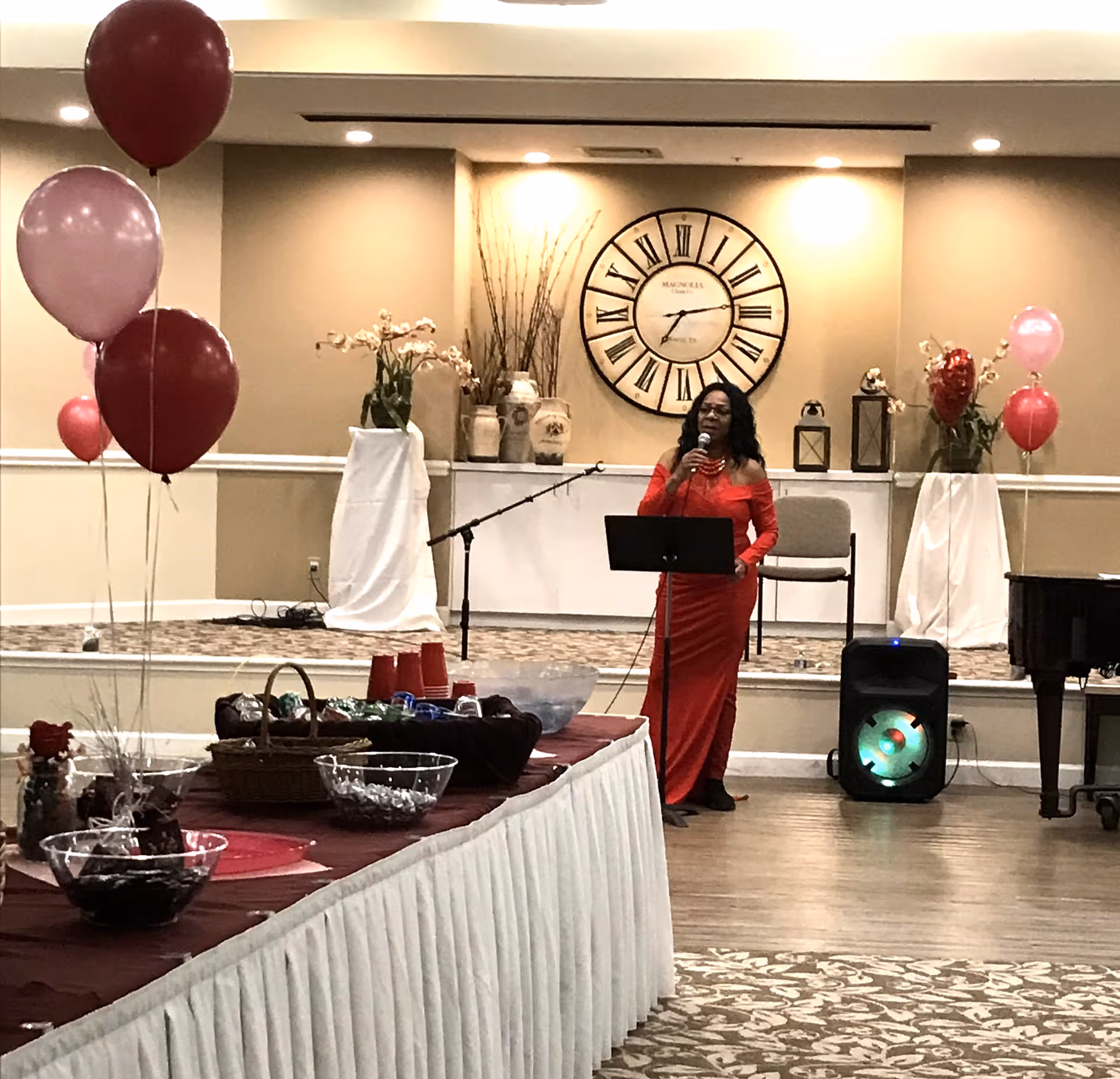 A woman in a red dress is speaking into a microphone in a decorated room. There are red and pink balloons on either side of the room, a large clock on the wall behind her, and a table in the foreground with bowls of snacks and red cups. The room has a patterned carpet and a speaker on the floor near the woman.