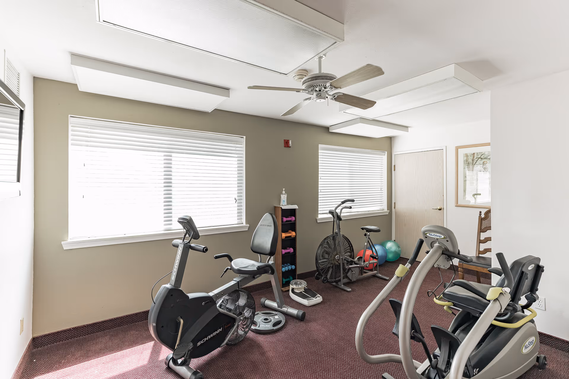 A small exercise room with stationary bikes, a recumbent bike, dumbbells on a rack, exercise balls, and a scale. The room has two windows with blinds, a ceiling fan, and a framed picture on the wall.