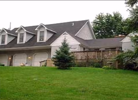 Exterior view of a residential-style building with a sloped roof, three dormer windows, a three-car garage, a wooden fence, and a green lawn with trees.
