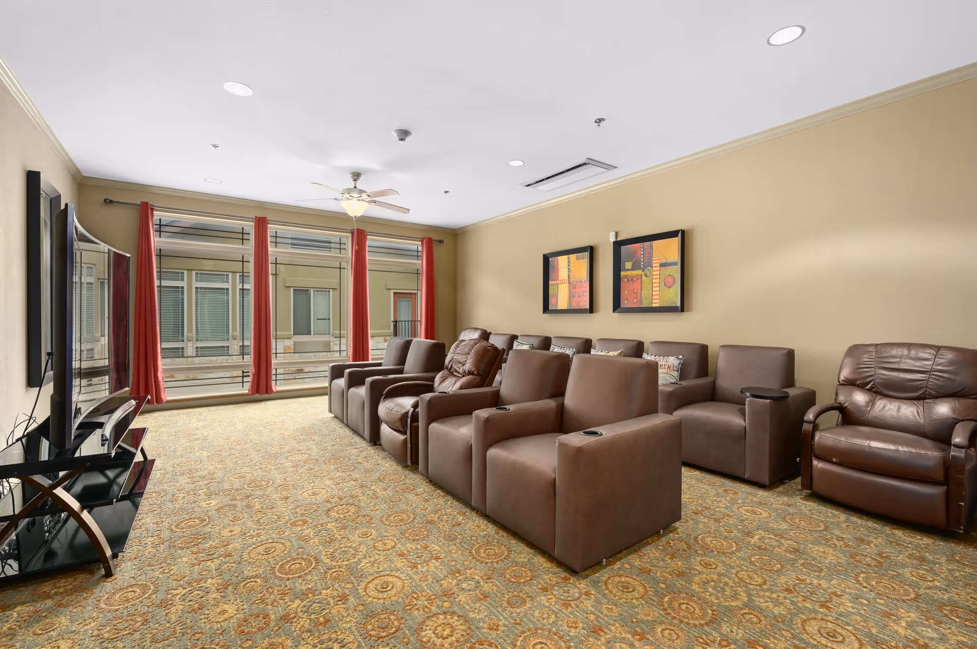 Media lounge with multiple brown recliner chairs facing a flat-screen TV and large windows with red curtains.