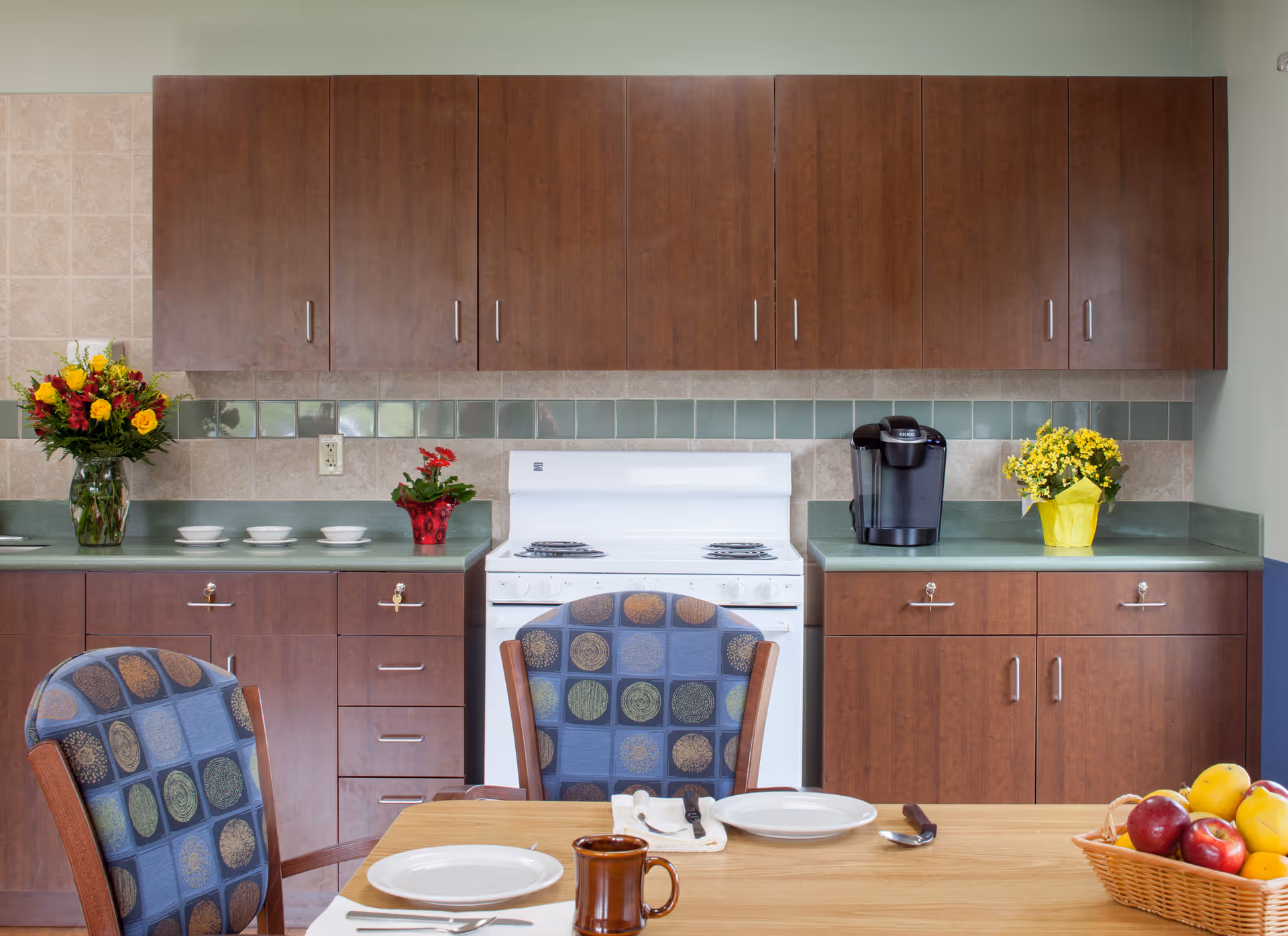 A kitchen area with wooden cabinets, a white stove, a coffee maker, and a countertop decorated with flower vases. In the foreground, a wooden dining table is set with plates, utensils, a mug, and a basket of fruit. Two chairs with patterned upholstery are partially visible.