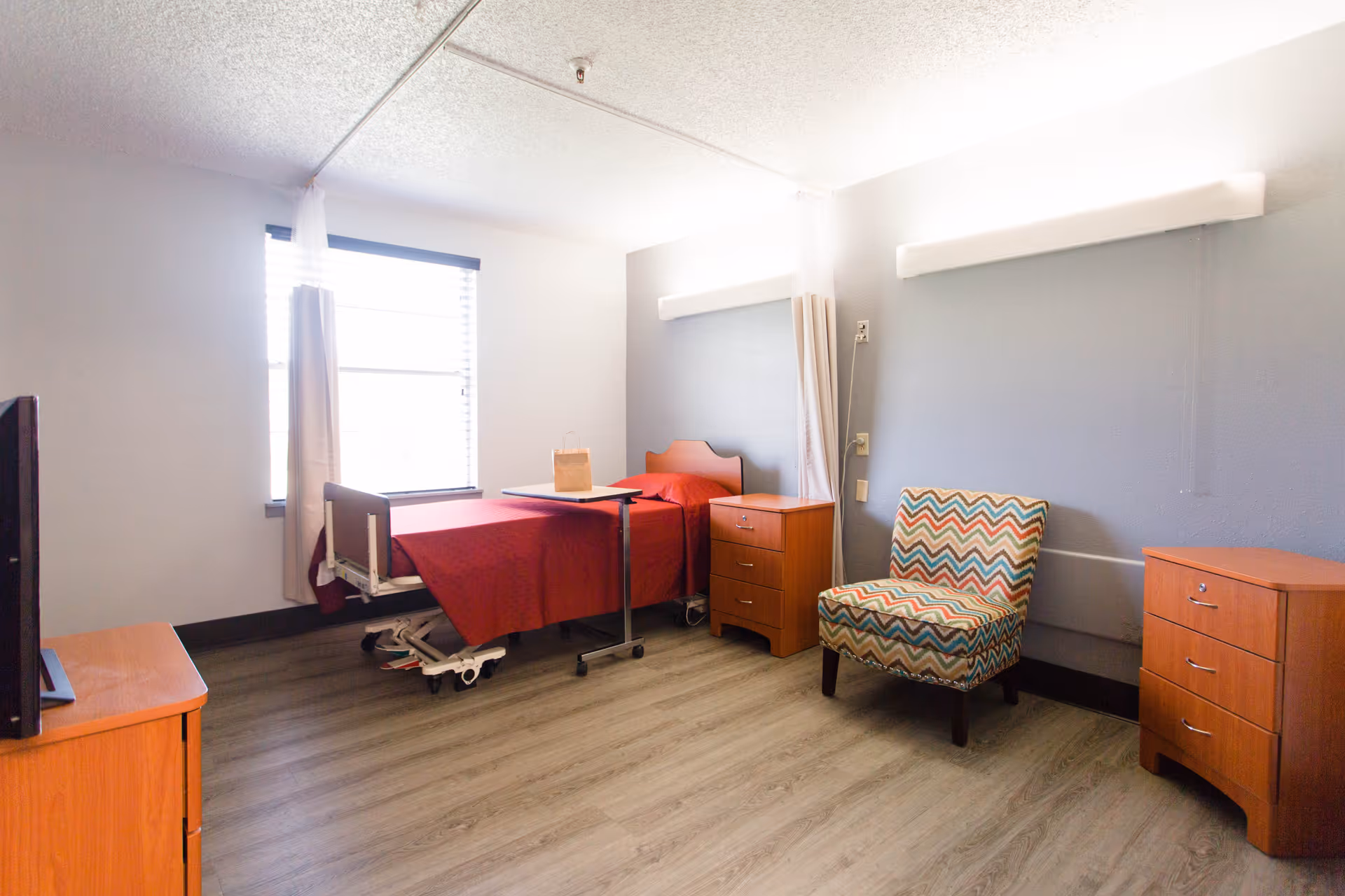 A simple, clean bedroom in a senior living facility with a single hospital-style bed covered in a red blanket, a small overbed table, two wooden nightstands, a colorful patterned armchair, a window with white curtains, and a TV on a wooden stand.