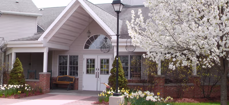 Entrance of a senior living facility with a covered porch, white double doors, brick pillars, a bench, a street lamp, and blooming white flowers on a tree and in the garden beds.