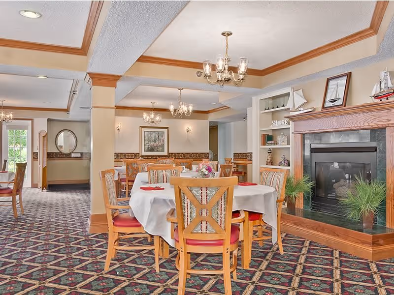 A bright and spacious dining room with multiple wooden tables and chairs arranged neatly. Each table is covered with a white tablecloth and has a small floral centerpiece. The room features a patterned carpet, beige walls with wooden trim, and several chandeliers hanging from the ceiling. There is a fireplace with a wooden mantle decorated with model sailboats and framed pictures. Built-in shelves next to the fireplace hold books and decorative items. Large windows allow natural light to fill the space.